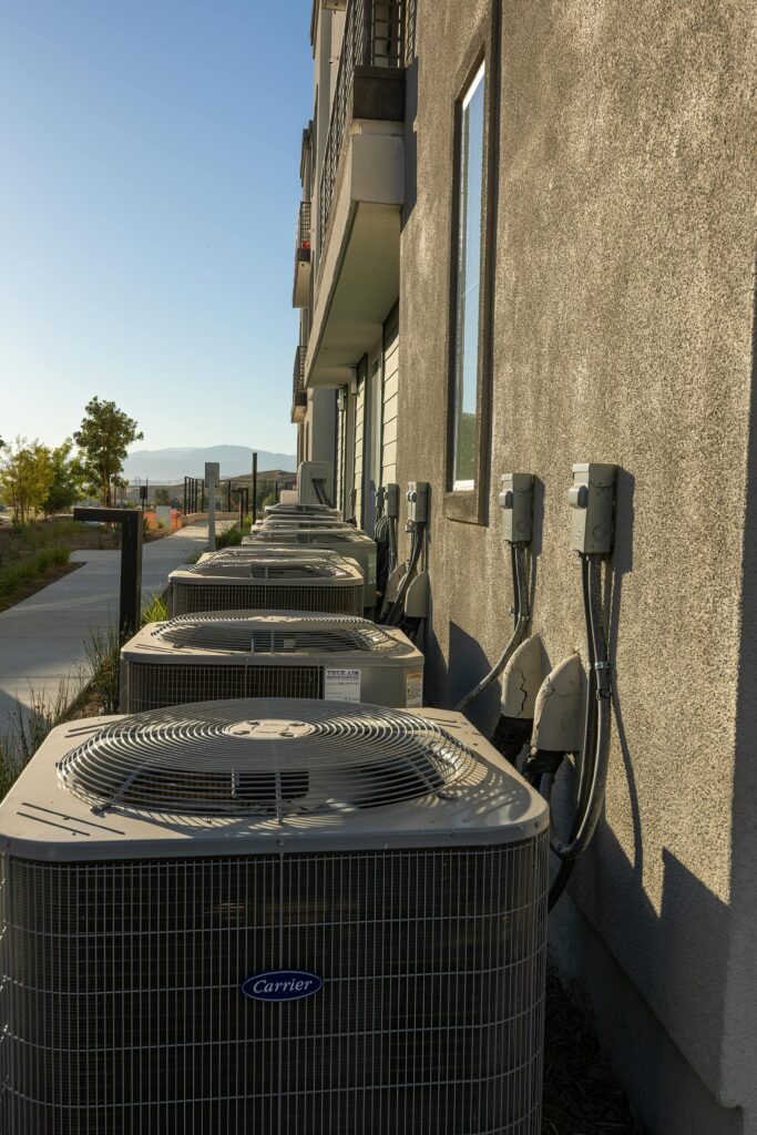 pexels-photo-17602538-17602538 Line of air conditioning units on a city apartment exterior with a sidewalk view.